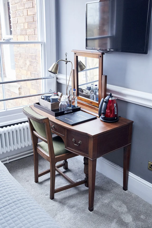 The image shows a serene bedroom with a wooden desk, a green chair, and a mirror above it. A lamp with a black shade and a red lamp are on the desk, along with a red coffee mug and a black notebook. A red and black alarm clock is on the desk, and a white radiator is on the floor. A window with a white curtain allows natural light to enter, and a black television is mounted on the wall. The room is well-organized and comfortable, with a harmonious color palette.