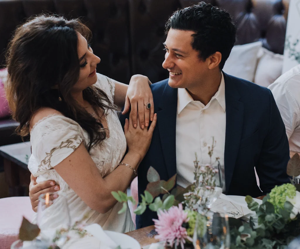 In the image, actor Benji Li is captured in a romantic moment with actress Emma Stone, seated at a pink table with a flower arrangement and wine glasses. Benji wears a white lace dress and Emma dons a navy blue suit. The scene is set in a cosy, intimate room with a pink couch and white wall.