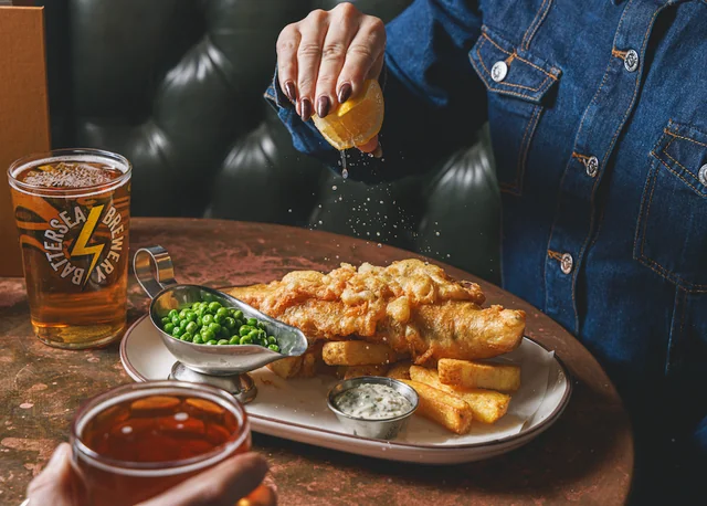 A woman in a blue denim jacket is enjoying a meal at a restaurant, with a plate of fried fish, fries, and green peas on a wooden tray. She is pouring condiments into a bowl and holding a silver spoon. Two glasses of beer, one with a yellow label and the other with a brown label, are on the table. The scene is set against a black leather couch and a brown tablecloth, creating a cozy atmosphere.