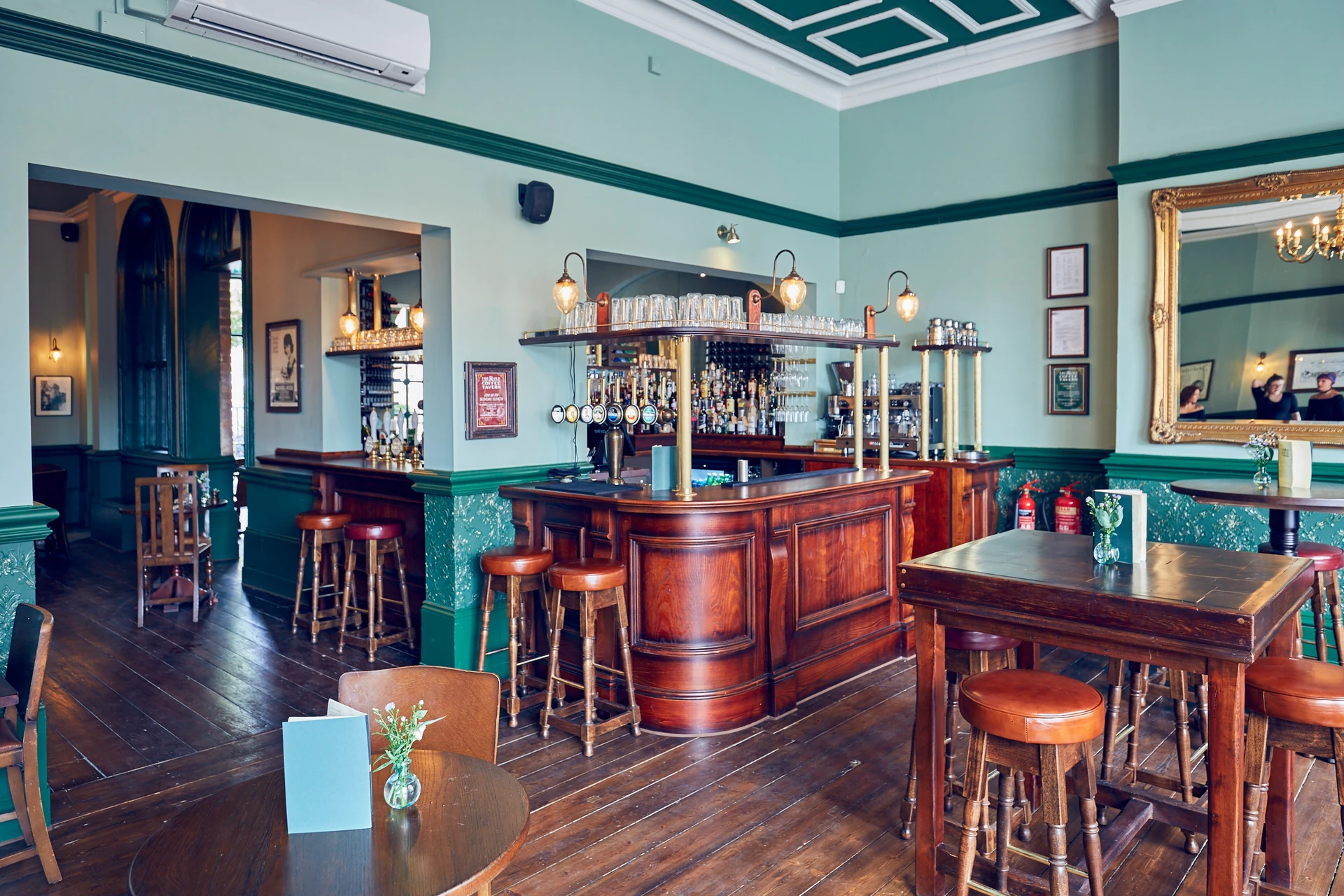 The image shows a cozy, well-lit bar with a wooden bar, six stools, and a mirror above. The walls are green, and the floor is wooden. A vase with a flower adds a touch of nature. The bar is equipped with a bar cart and a mirror. The scene is inviting and comfortable, with a warm atmosphere.
