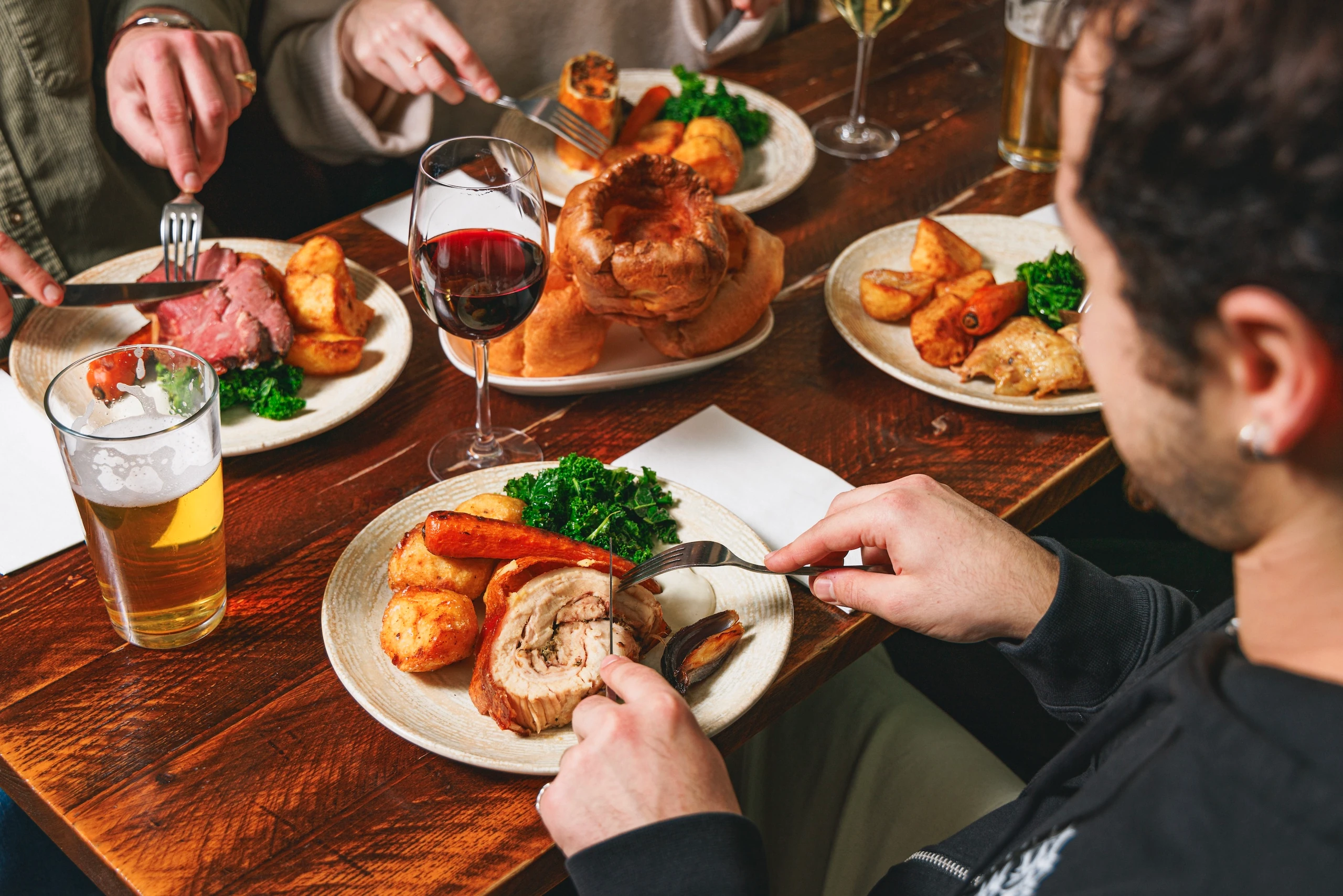A cozy dining scene features a wooden table with various dishes, including a large roast, roasted potatoes, and a salad. Two people are present, one with a silver fork and knife, and another with a silver knife and fork. A glass of beer and a wine glass are also present. The atmosphere is warm and inviting, with a focus on the food and company.