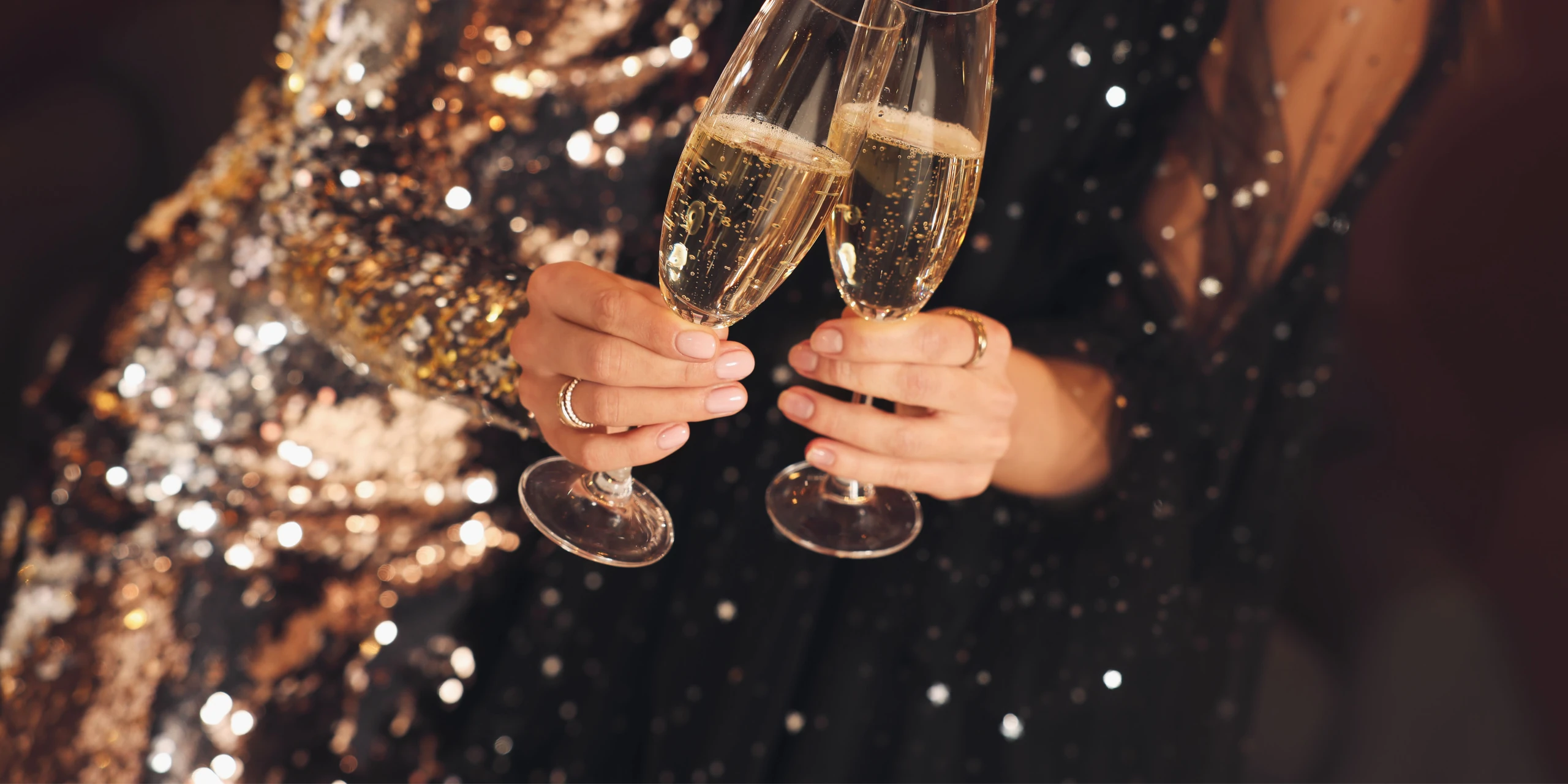 A pair of champagne glasses, filled with bubbly champagne, are held by hands in the center of the image. The background is a dark, shimmering surface, and the focus is on the glasses and hands, creating a celebratory atmosphere.