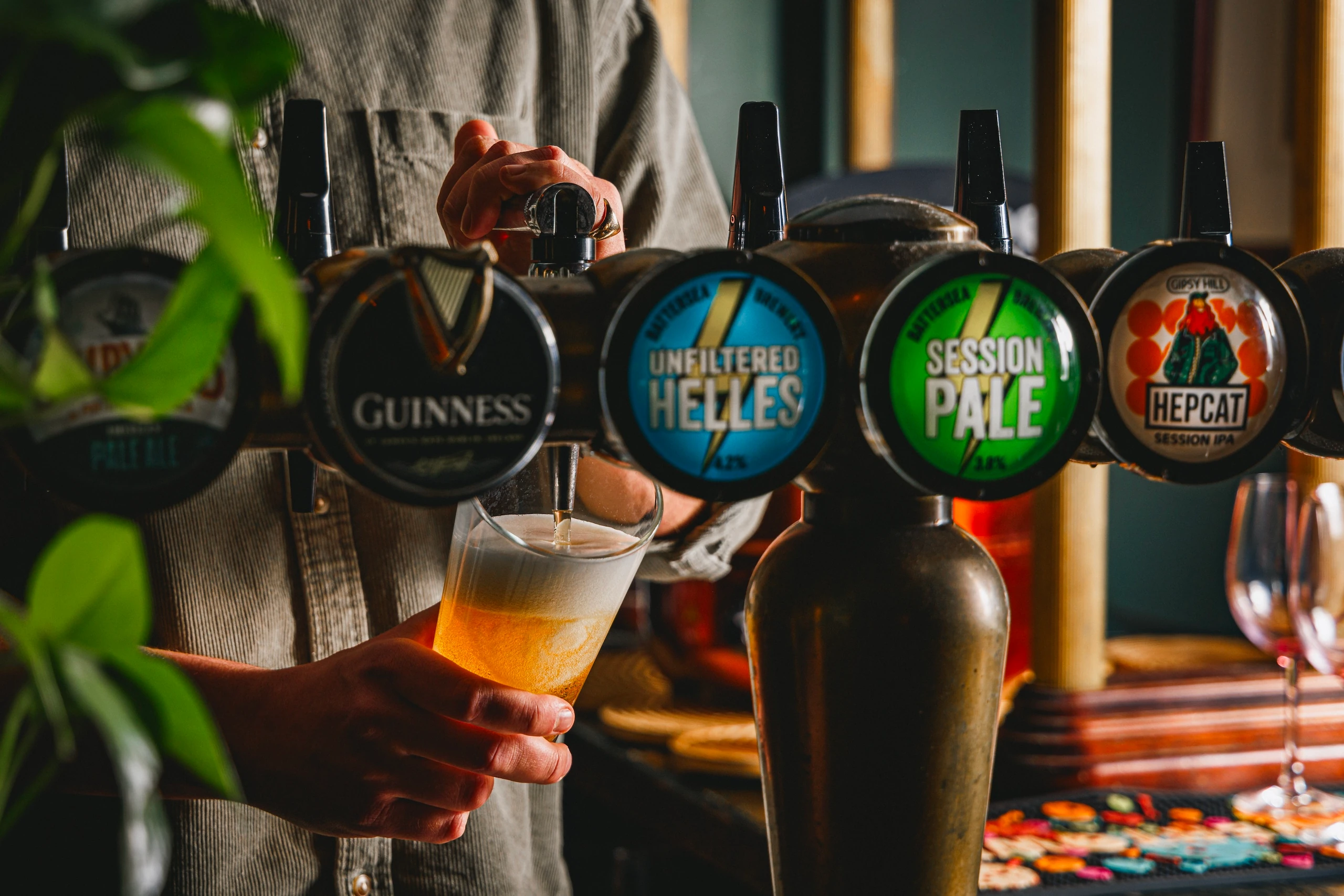 A person in a gray shirt is pouring beer from a row of tapbells in a bar, with a frothy beer in a glass. The tapbells display various colors and logos, and the background features a wooden counter and green plant. The scene captures a moment of anticipation and enjoyment in a bar setting.