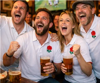 In a lively bar, four friends in white polo shirts with red roses on their sleeves celebrate. They hold beer glasses filled with beer, laughing and enjoying the moment. A television screen in the background shows a soccer match, adding to the festive atmosphere.