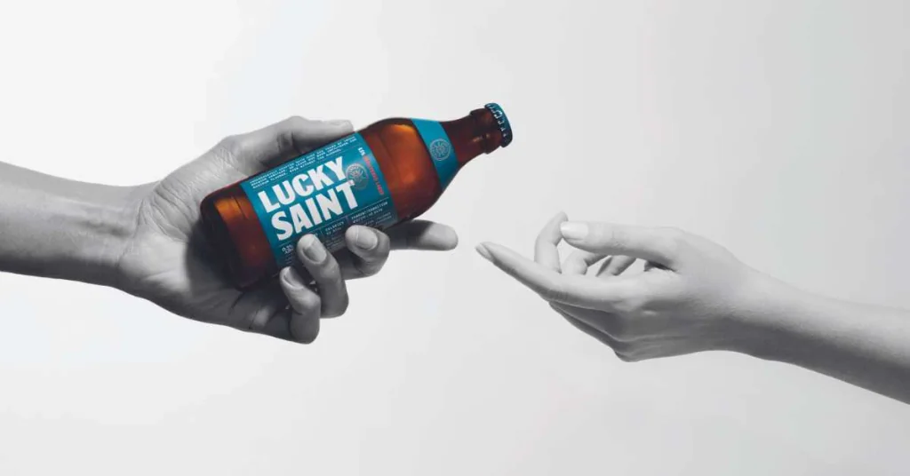 A hand holds a brown beer bottle with a blue label, inviting another hand to receive it. The hand is positioned to the right, and the beer bottle is on the left. The background is white, emphasizing the objects and their actions. The image is in black and white, emphasizing the colors and details.