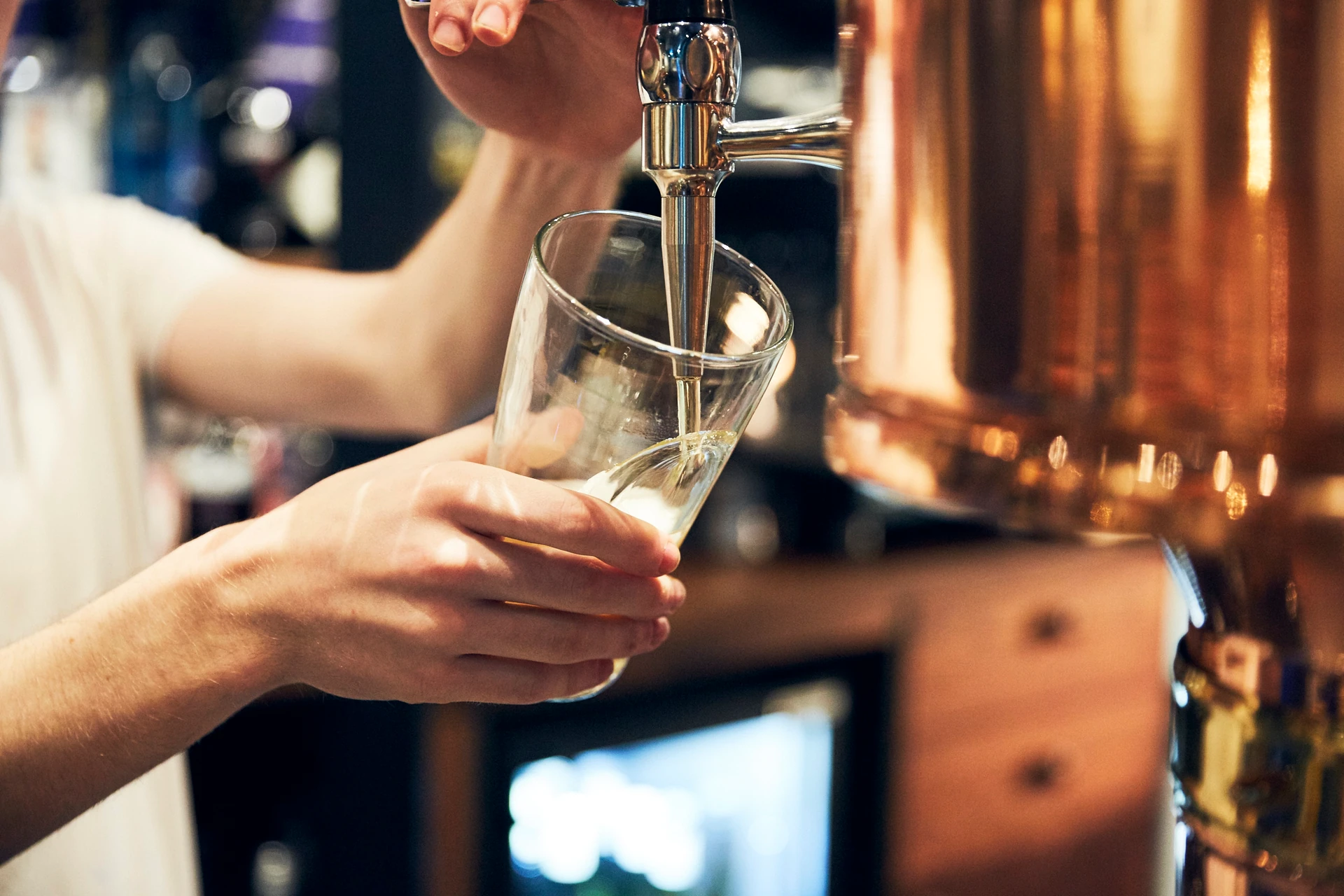 A person in a white shirt is pouring a frothy, frothy white liquid into a clear glass with a silver spout, using a copper-colored glass distillation machine. The background is blurred, suggesting a bar or restaurant setting. The image captures a moment of skill and precision in the craft of distilling.