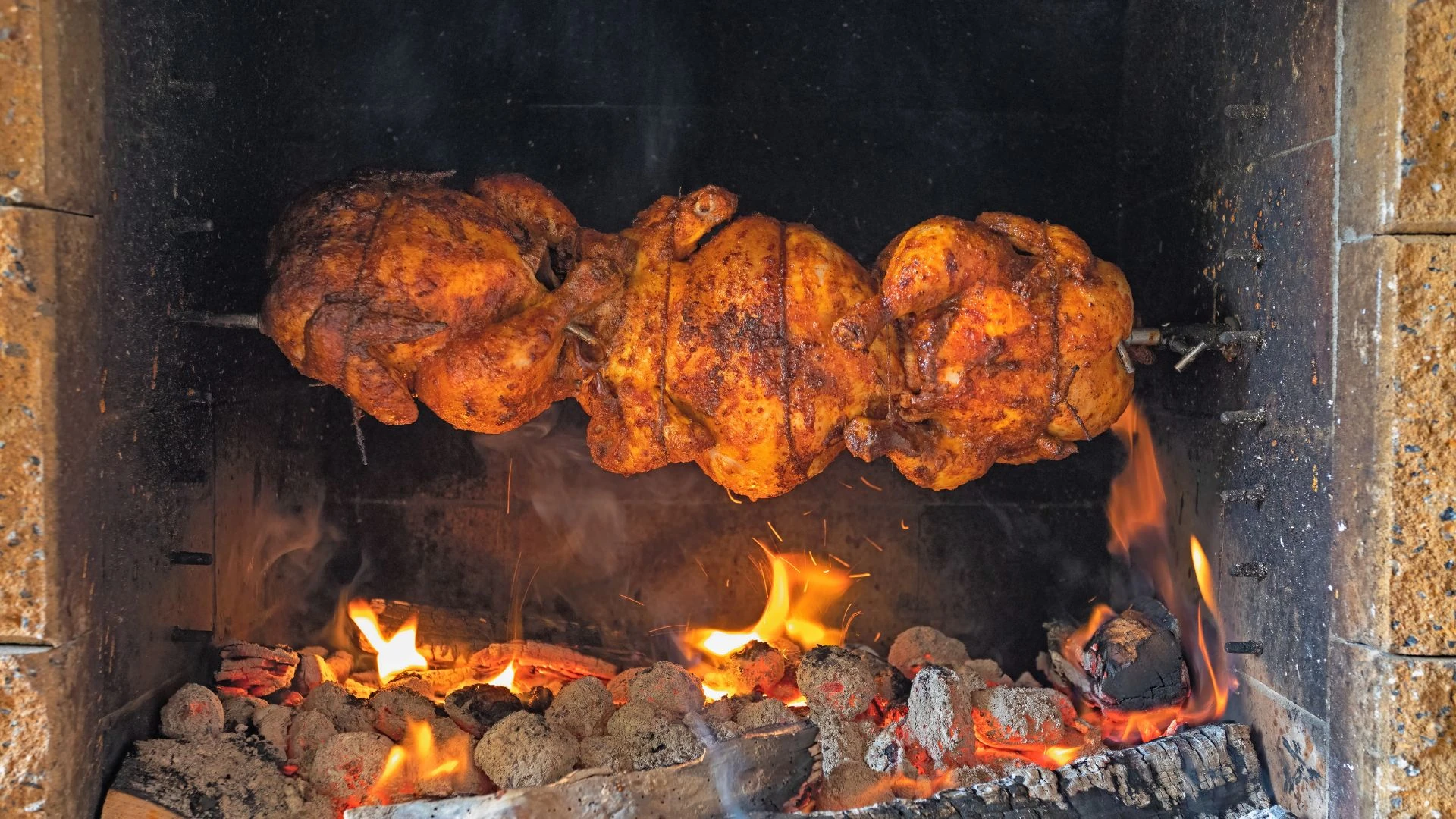 A large, black oven with a glass door is the main focus of the image, filled with two roasted chickens. The chickens are golden-brown and are being cooked over a fire, with flames dancing around them. The oven is surrounded by a pile of firewood, and the scene is set against a dark brick wall. The image captures a moment of culinary artistry and warmth.