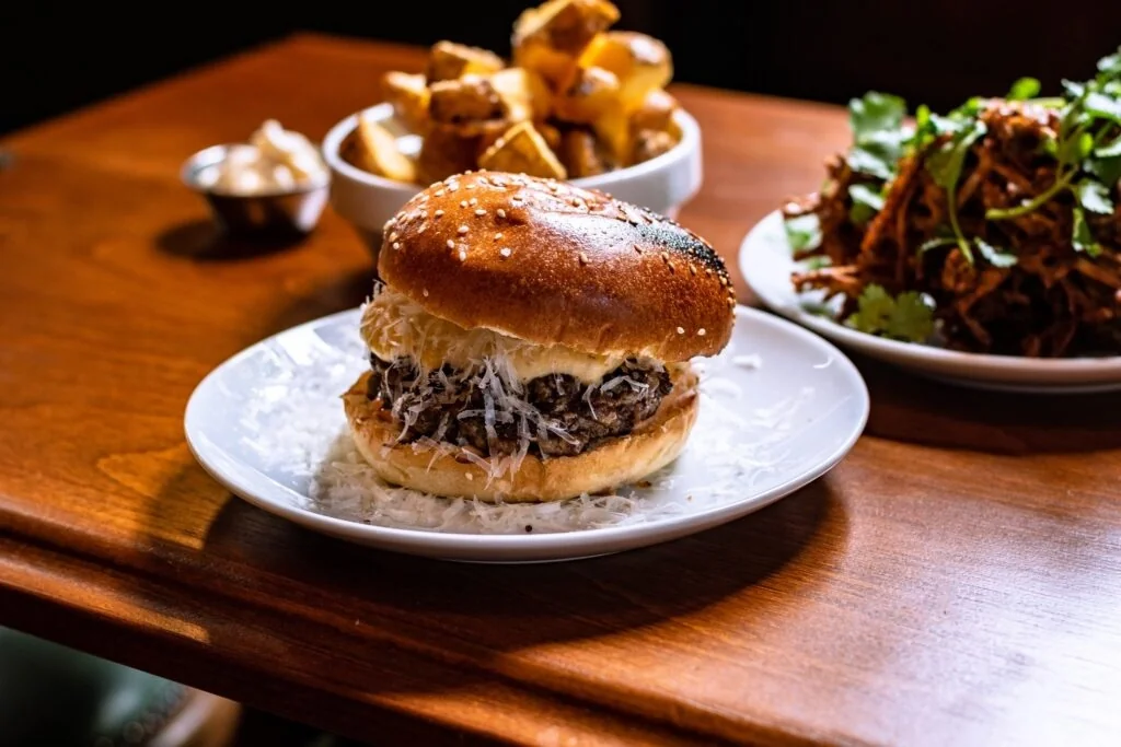 A wooden table features a large, golden-brown burger with sesame seeds, topped with shredded meat and melted cheese, accompanied by a side of crispy fries and a small bowl of fresh parsley salad. The scene is set against a dark background, emphasizing the vibrant colors and textures of the food.