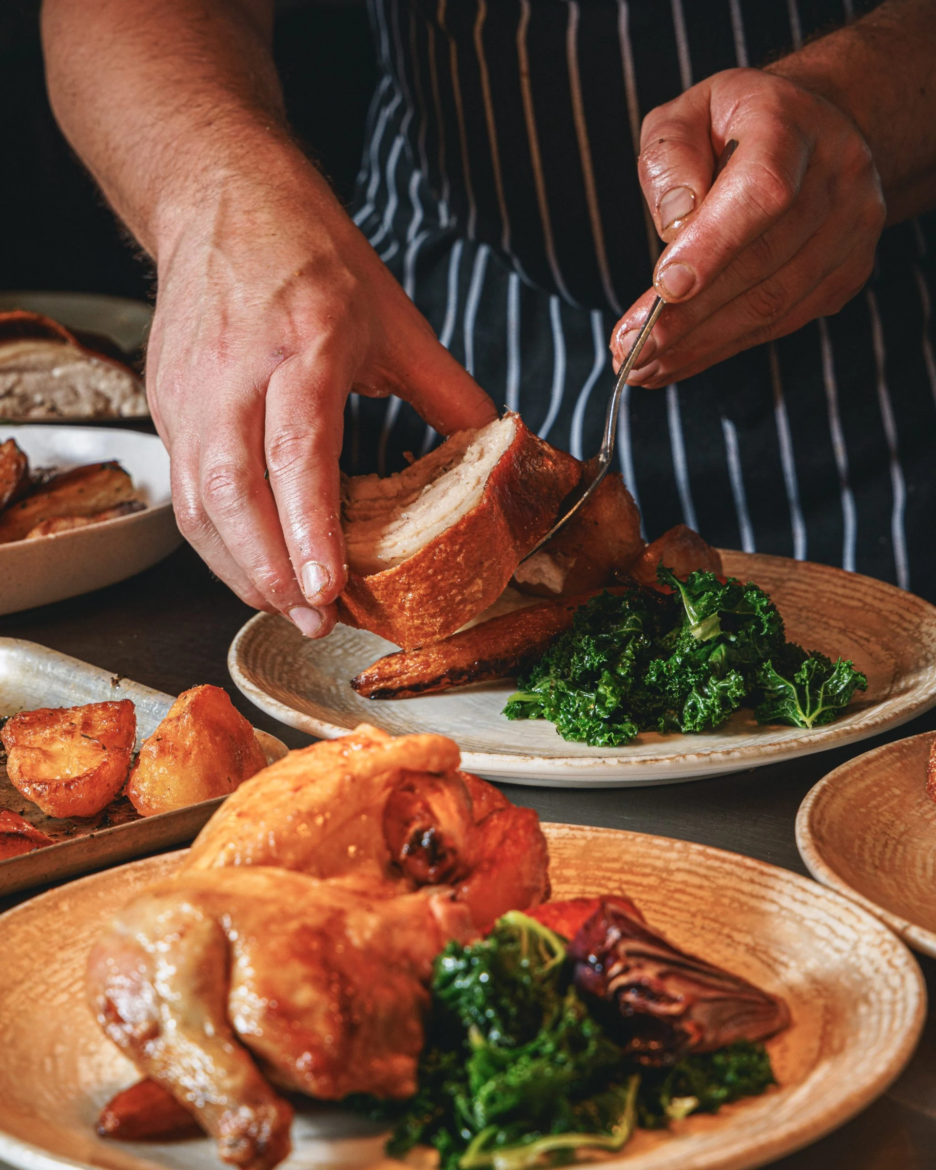 A person in a striped apron is preparing a meal, with a golden-brown chicken breast on a plate and a piece of bread on another. The chicken is being cut with a knife, while the bread is on a plate with green vegetables. The scene takes place on a wooden table, with a dark background emphasising the colours and textures.