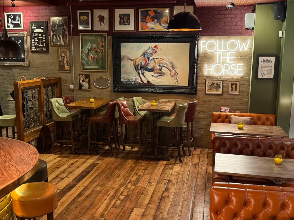 The image shows a cozy, rustic restaurant with a wooden floor, brick walls, and a large, colorful painting of a horse. The walls are adorned with various paintings, including a large one with a horse. The restaurant features wooden booths, leather booths, and a bar with a red leather booth. The lighting is warm and inviting, with a neon sign reading "RIDE ON THE Horses" and a hanging lamp. The atmosphere is cozy and welcoming, perfect for a horse-themed restaurant.