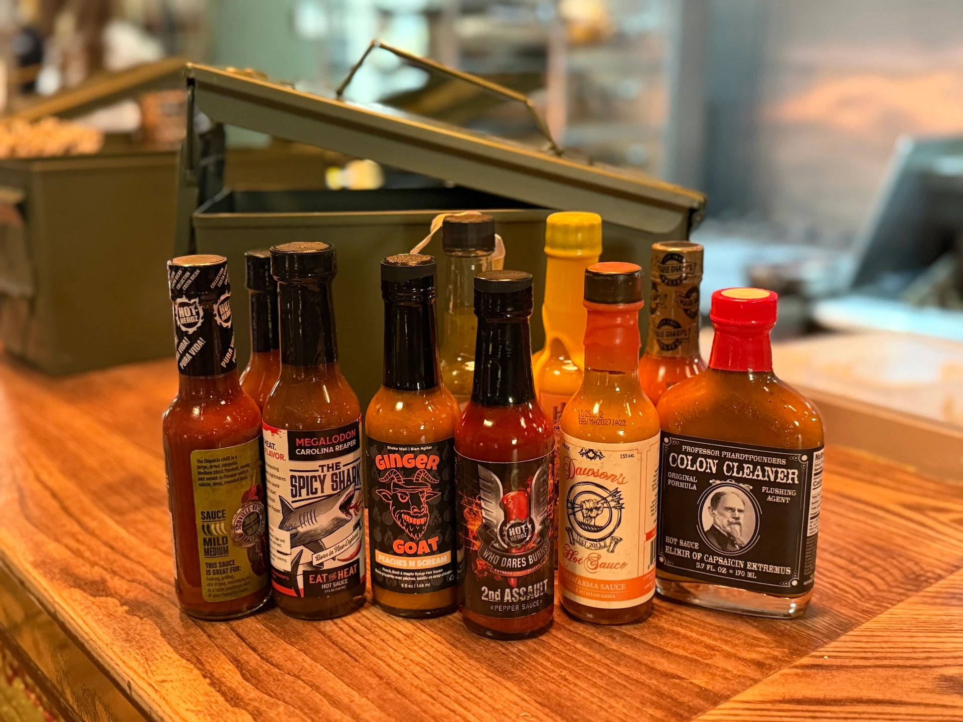 A wooden counter displays nine unique condiment bottles, each with a distinct label and cap. The bottles vary in color and size, with some featuring images of animals. A green container is in the background, and the scene suggests a store or kitchen setting.