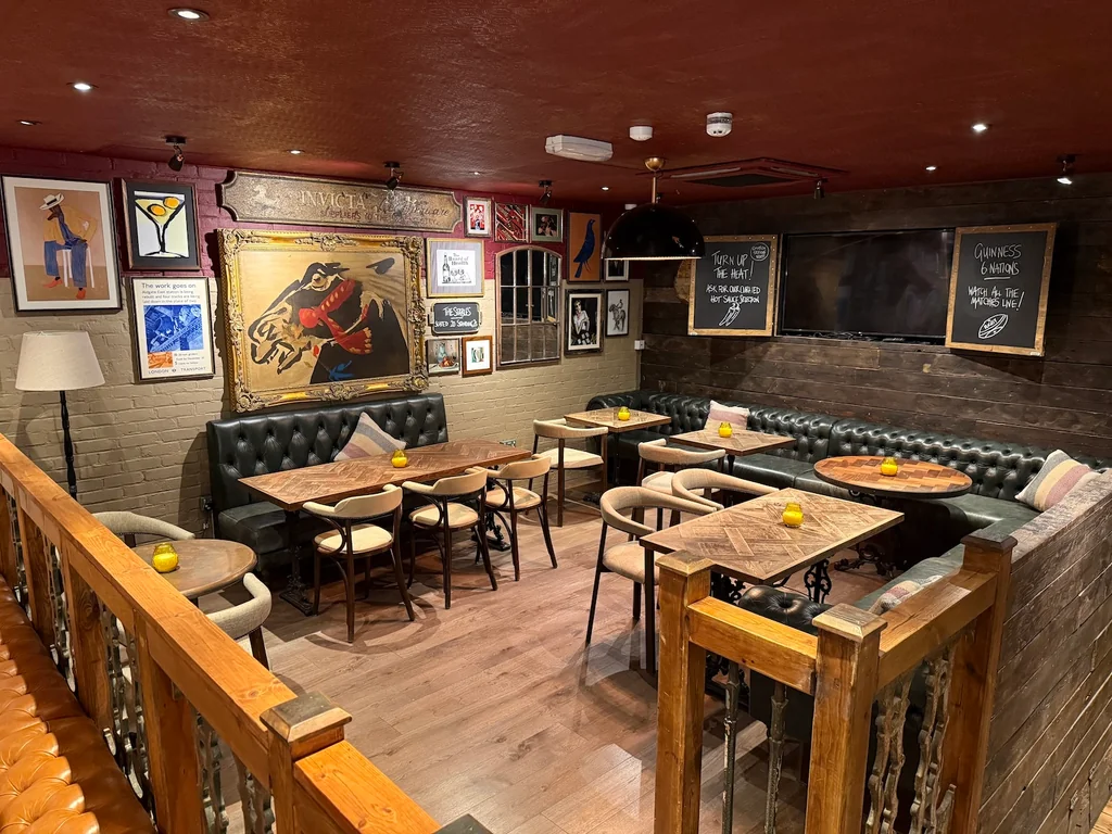 The image shows a cozy, rustic restaurant with dark wood floors and a brick wall. The walls are adorned with various paintings and posters, and a chalkboard menu is displayed on the wall. Tables and chairs are arranged for patrons, and a wooden bar is present for drinks. The atmosphere is warm and inviting, with a cozy ambiance.