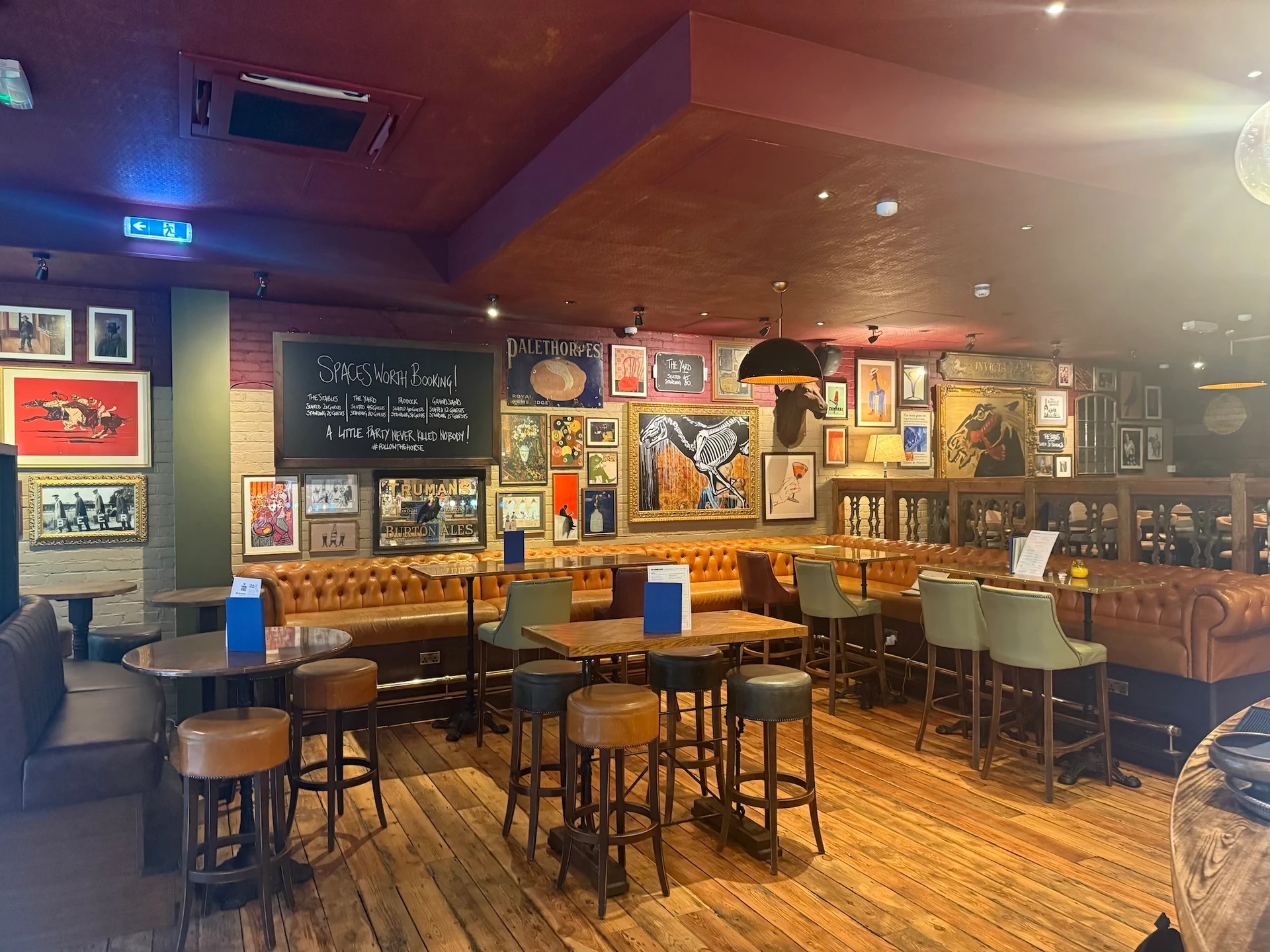 The image shows a cozy, rustic bar with wooden floors and walls, featuring a bar counter with blue and white bar stools, a black chalkboard menu, and a wooden shelf with various items. The walls are adorned with framed pictures and paintings, and the ceiling has a black ceiling light. The bar is well-lit, creating a warm and inviting atmosphere.
