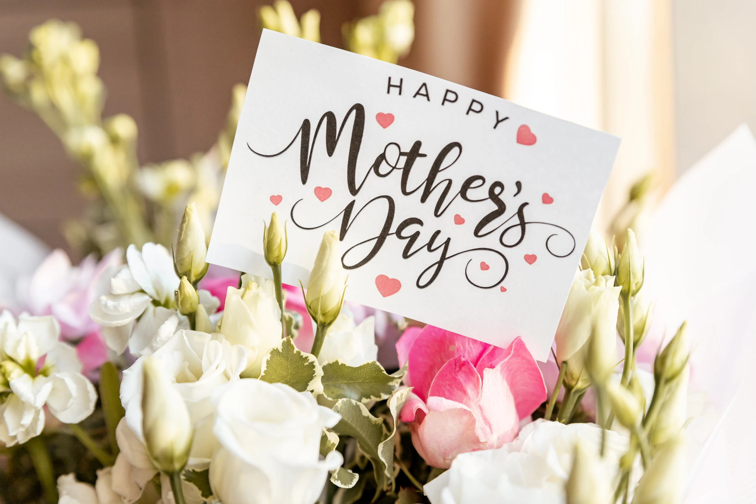 A bouquet of white and pink roses, with green leaves, is the main focus of the image, placed on a white card with a pink heart and the message "Happy Mother's Day" in black cursive font. The card is placed on a white tablecloth, with a window in the background. The arrangement creates a warm and inviting atmosphere.
