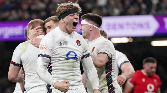 A rugby player in a white jersey with red and blue stripes celebrates a goal in a stadium filled with spectators. His teammates, also in white jerseys, surround him, with some holding hands and others cheering. The image captures the excitement and camaraderie of the sport.