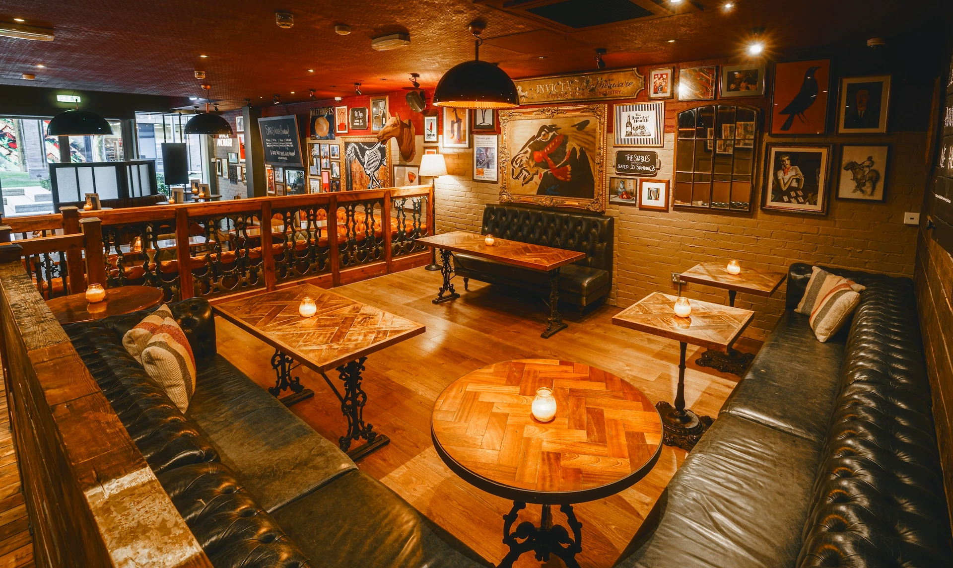 The image shows a cozy, rustic bar with wooden floors and walls, featuring a wooden bar, a wooden table with candles, and a black chandelier. The bar has a wooden counter and a blackboard menu. The walls are adorned with framed pictures and a blackboard menu. The bar is filled with patrons, creating a warm and inviting atmosphere.