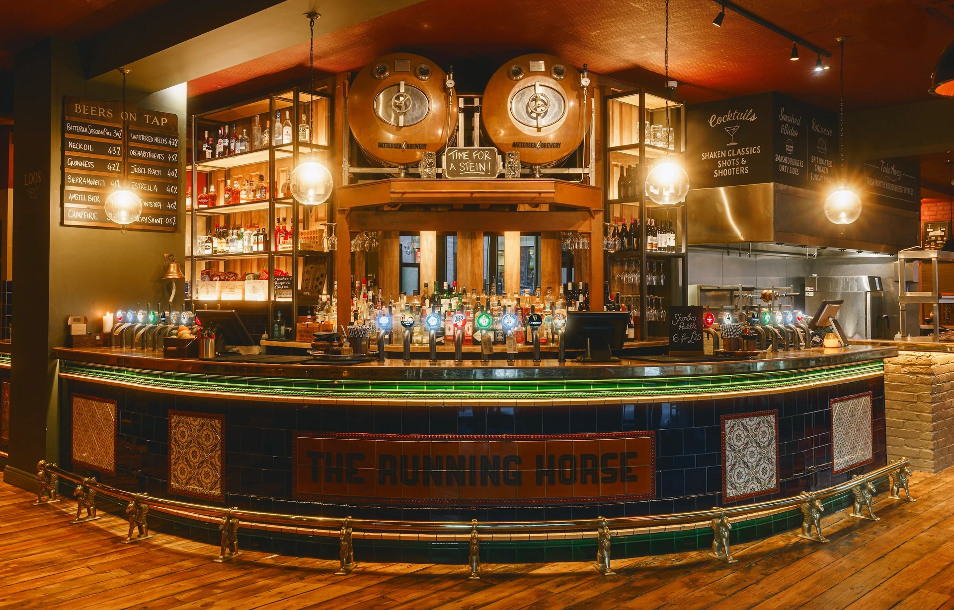 The image shows a well-lit, cozy bar with a green-tiled counter, a large wooden bar, and a metal railing. The counter has a large shelf of liquor bottles and a large clock. The bar is surrounded by a blackboard menu, a black sign, and a black metal bar. The floor is wooden, and the ceiling has a black and white chalkboard menu. The atmosphere is warm and inviting, with a sense of order and organization.