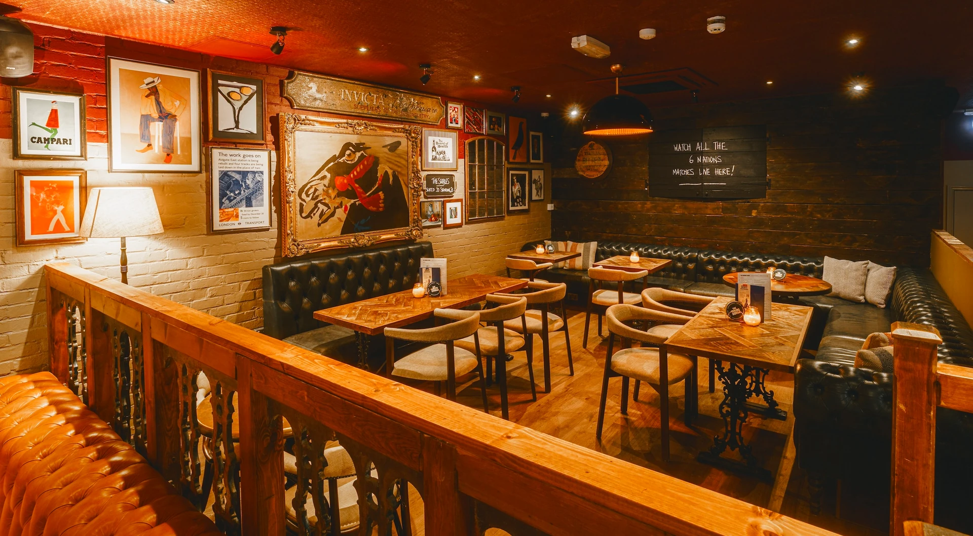 The image shows a cozy, rustic restaurant with dark wood floors and brick walls, featuring wooden furniture, a wooden bar, and a wooden railing. The walls are adorned with framed pictures and a large blackboard with a "Welcome to the pub" sign. The ceiling has hanging lights and a vent. The atmosphere is warm and inviting, with a cozy seating area and a fireplace for warmth.