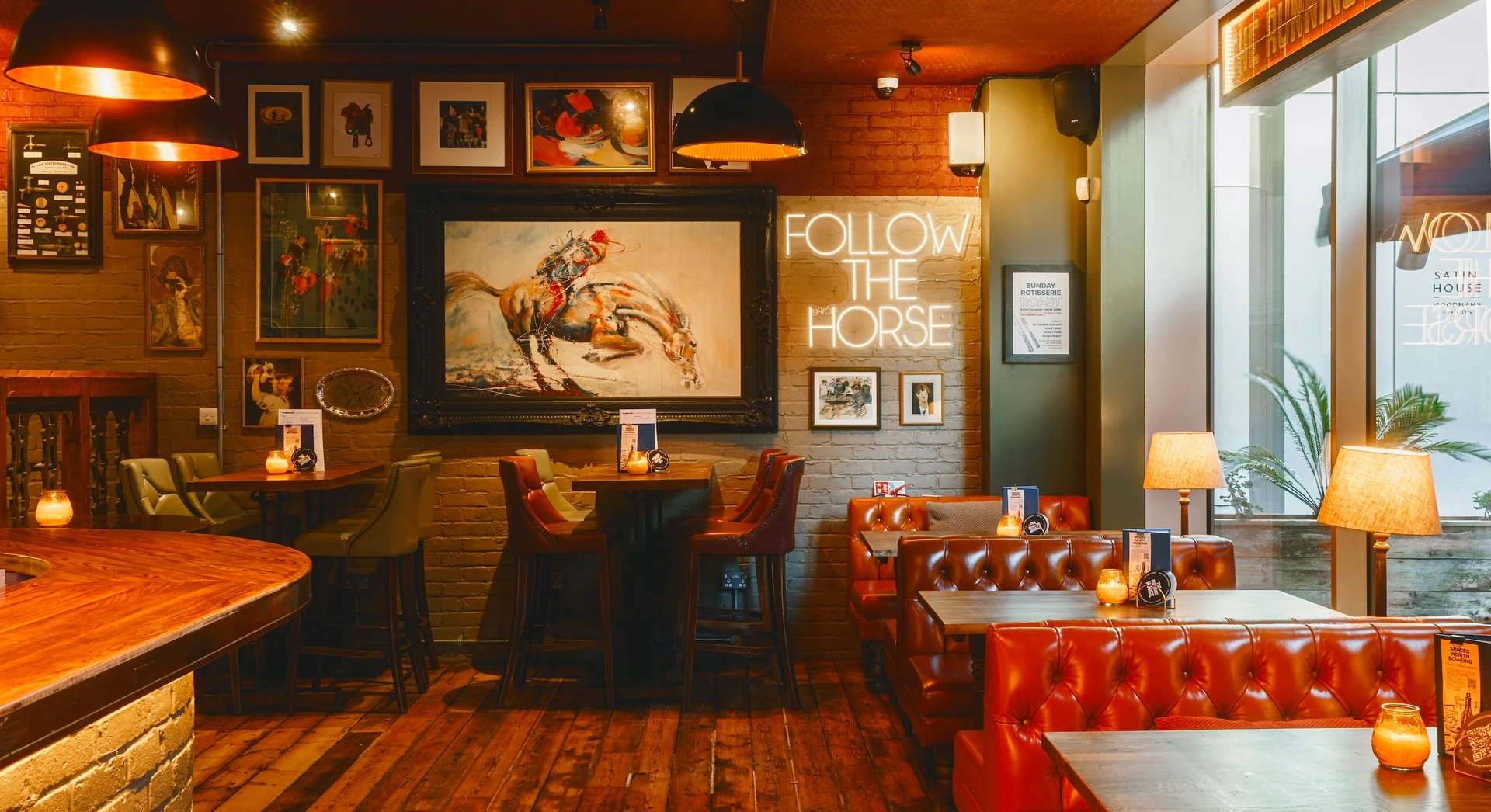 The image shows a cozy, rustic bar with wooden furniture, a bar counter, and a large window. The walls are adorned with various paintings and signs, and the floor is covered in a warm, earthy color. The bar is equipped with a barstool and a large mirror, and the overall atmosphere is inviting and warm.