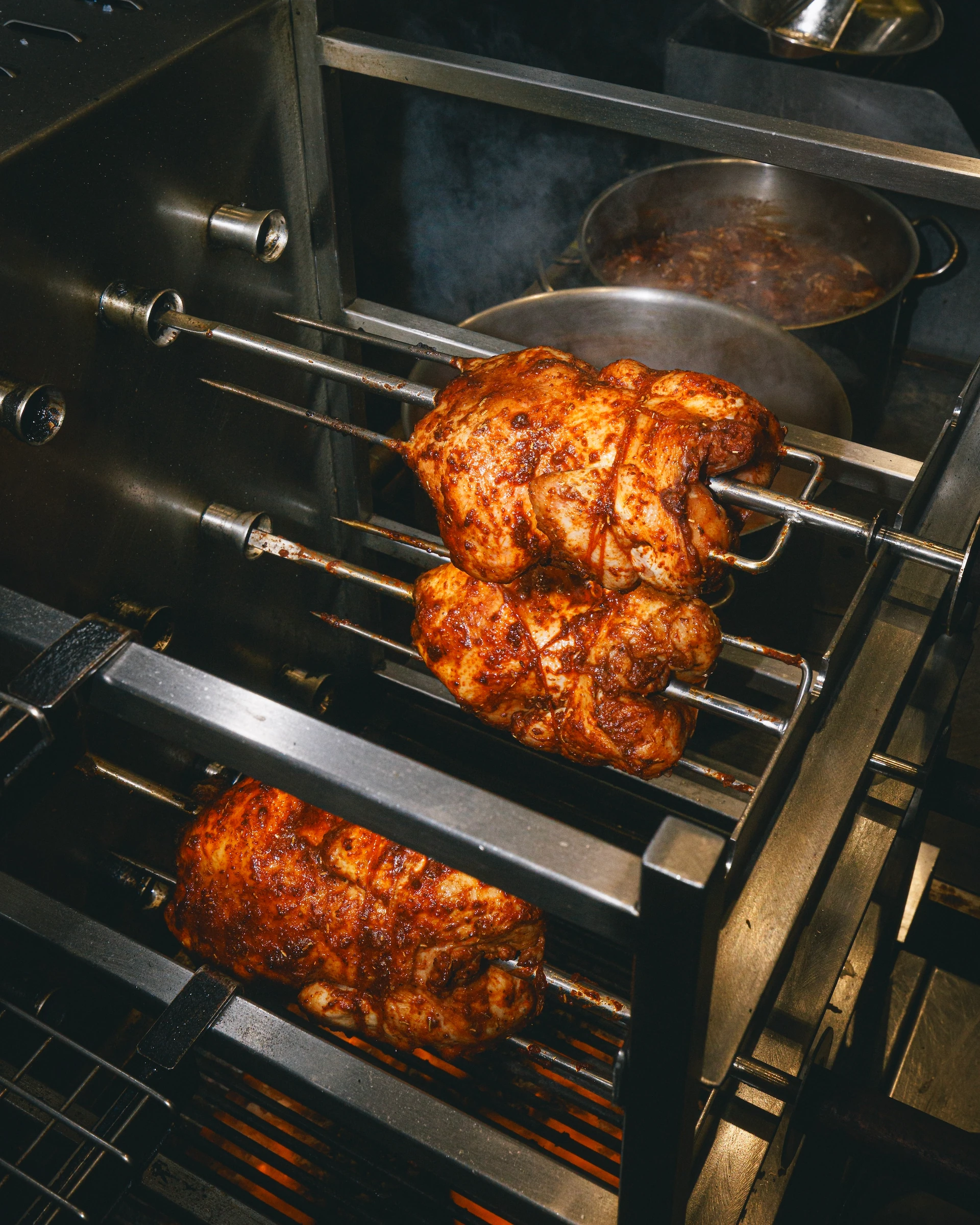 In a kitchen, a large, silver oven with two metal racks is in use, cooking two pieces of meat on the upper rack. The meat is golden brown and has a crispy texture. A pot of sauce is nearby, and the oven has a metal handle and knobs. The scene is set against a dark background, emphasizing the oven and its contents.