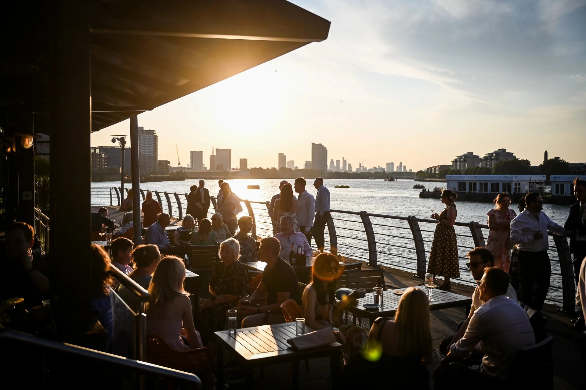 At a rooftop bar in Singapore, people enjoy the sunset view of the Singapore River and skyline. The bar is surrounded by a metal railing and wooden tables, with a large awning providing shade. The scene is bathed in warm, golden light, creating a serene atmosphere.
