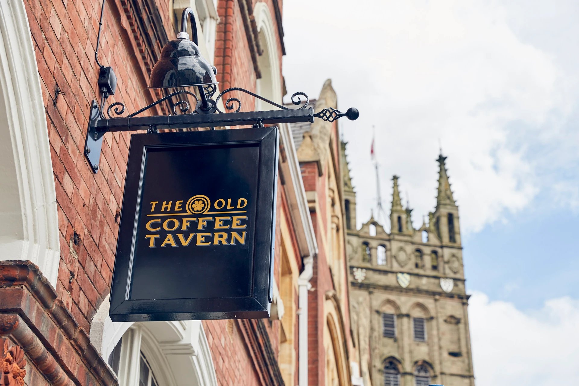 The image features a brick building with a black signboard displaying "THE GROVE CAFE" and "Cafe 100". A black lamp post is attached to the signboard, and a Gothic spire is visible in the background. The scene is set against a clear blue sky with white clouds, creating a serene atmosphere.