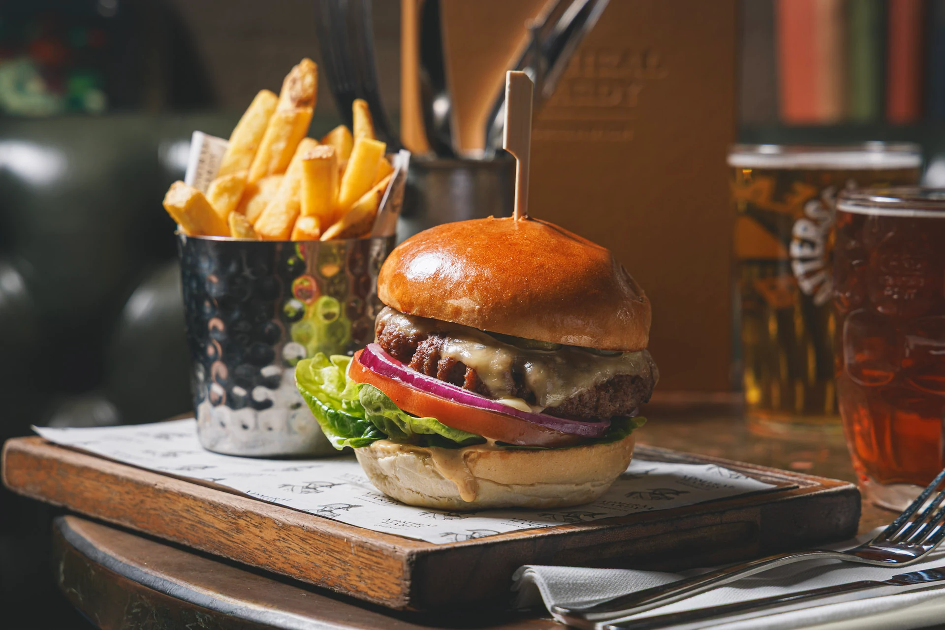 A wooden table features a large, well-prepared burger with a bun, lettuce, tomato, and cheese, topped with a toothpick. A side of fries is to the right, and two glasses of beer are on the left. The scene is set in a cozy restaurant, with a menu and a napkin nearby. The image captures a moment of anticipation before a meal.