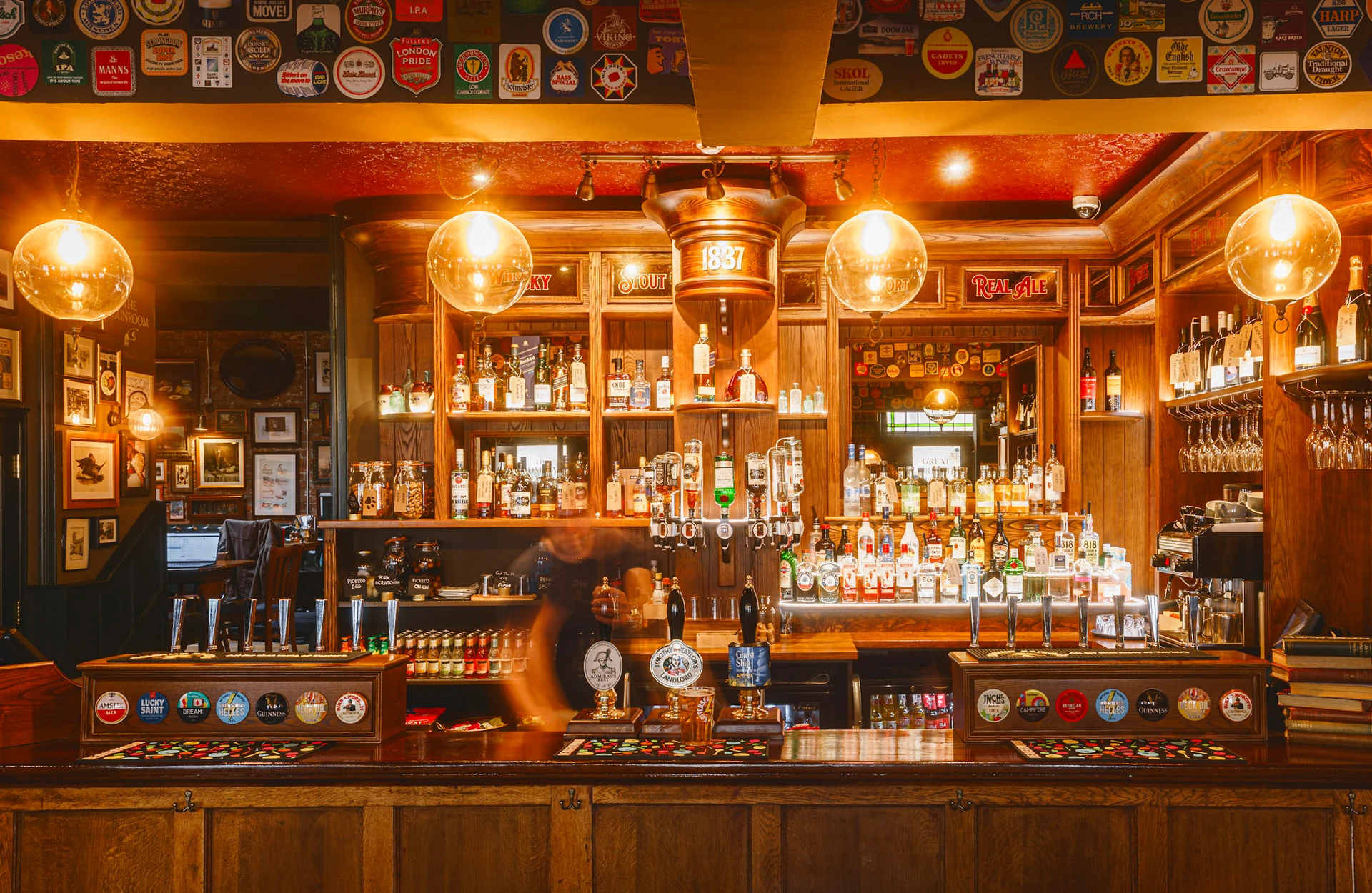 The image shows a cozy bar with a wooden counter, illuminated by warm lamps, featuring a variety of liquor bottles and glasses. The counter is adorned with framed pictures and a clock, and the bar is well-stocked with alcohol bottles. A bartender is present, and the atmosphere is inviting and relaxed.