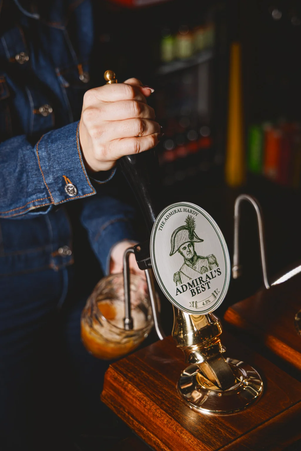 A person in a denim overalls is pouring a cold beer from a "Greenhouse" tap into a glass at a wooden bar. The background is blurred, suggesting a well-lit bar or restaurant. The image captures a moment of skill and precision in the art of beer brewing.