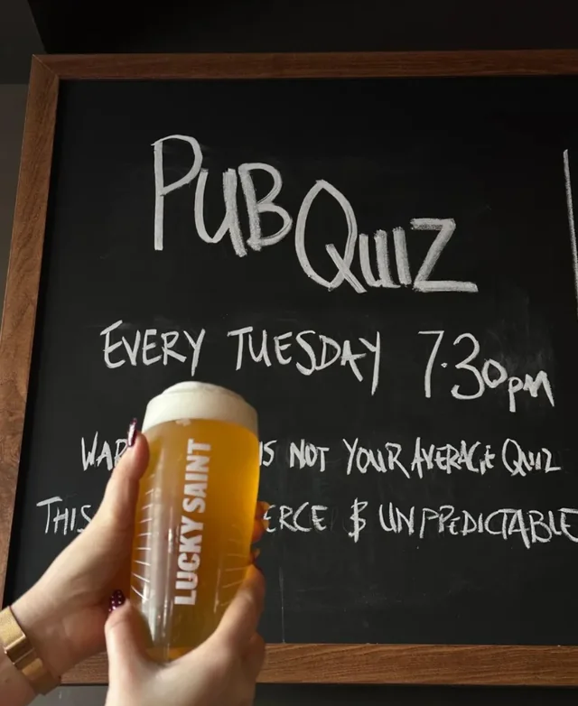 A person holds a pint of beer in front of a chalkboard menu for the "Puffin Tabbie" pub, featuring a 7:30am to 10:30pm shift, 11:30am to 12:30pm shift, and 1:30pm to 6:30pm shift. The menu lists drinks, specials, and specials for the day. The person's hand is visible, and the image captures the pub's atmosphere and menu.