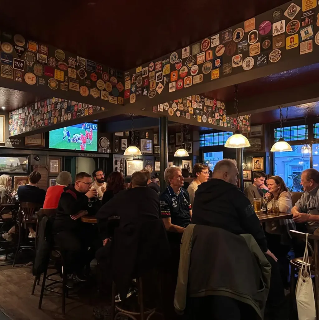 In a lively bar, people gather around tables, enjoying drinks and conversation. The bar is adorned with colorful posters and plaques, creating a vibrant atmosphere. A television screen displays a game, adding to the lively ambiance. The scene captures a moment of camaraderie and enjoyment in a bustling bar.