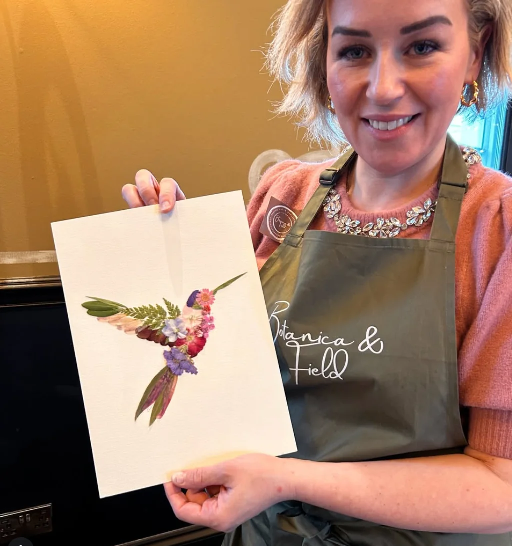 A woman in a pink sweater and apron, with a smile on her face, holds a watercolor painting of a hummingbird on a white paper. The painting features a vibrant mix of green leaves, purple flowers, and red berries. The woman is in a kitchen, with a stove and oven visible in the background. The painting is the main focus, with the woman and her artwork being the main subjects.