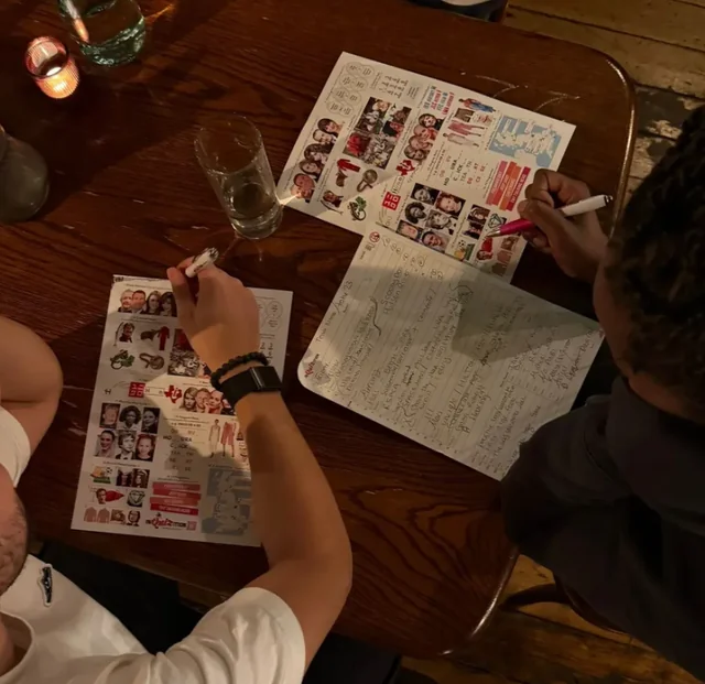 In a dimly lit room, two individuals are seated at a wooden table, engrossed in a creative activity. They are writing on paper with pens, surrounded by a variety of stickers and notes. A glass of water and a candle add to the cozy atmosphere. The scene captures a moment of collaboration and creativity in a shared space.