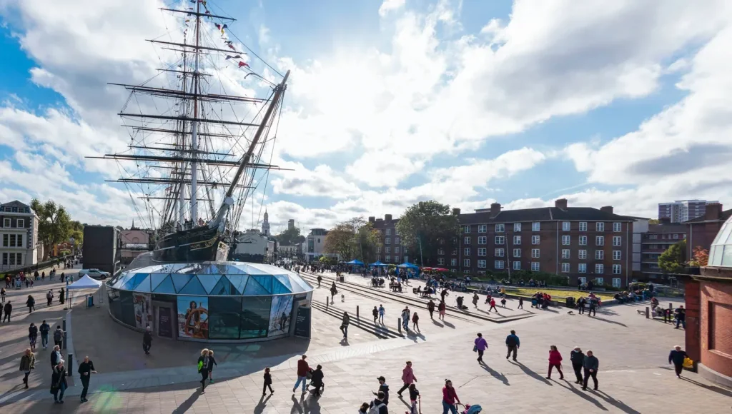 A large, three-masted tall ship is the centerpiece of a bustling square, surrounded by a glass dome and a red brick building. The square is filled with people, creating a lively atmosphere. The sky is a clear blue with scattered clouds, and the scene is set against a backdrop of trees and buildings. The image captures a typical day in a bustling city square.