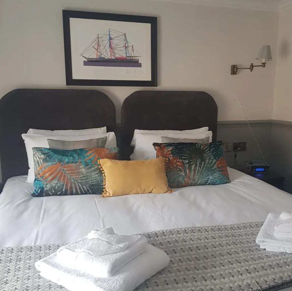 The image shows a well-organized bedroom with a large bed, adorned with colorful pillows and a white comforter. Two nightstands with lamps are on either side of the bed, and a painting of a ship hangs above it. The room is well-lit, creating a cozy atmosphere.