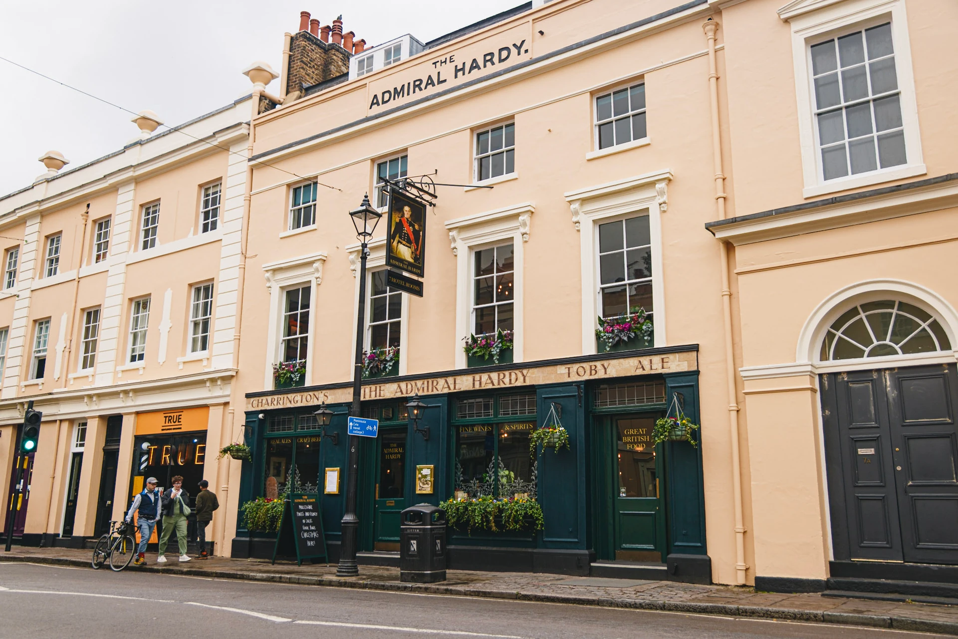 The image features the historic "The Old Bar & Barrels" pub in London, England, with a two-story building painted in warm yellow. The pub has a black awning and a green sign with the words "The Old Bar & Barrels". The street is bustling with people, and a bicycle is parked in front. The scene captures the lively atmosphere of the pub and its surroundings.