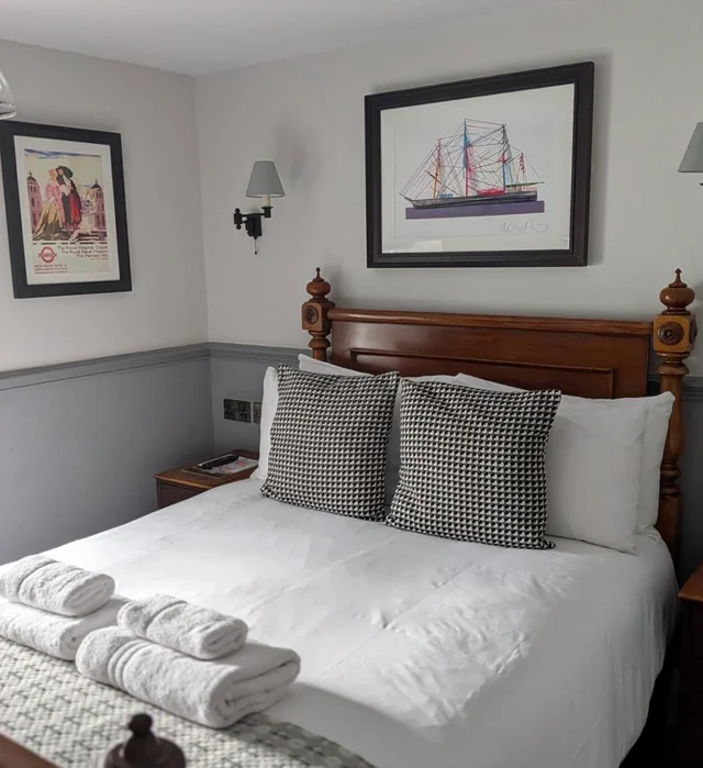 The image shows a serene bedroom with a large bed, white linens, and black and white checkered pillows. Two framed paintings hang above the bed, one of a ship and the other a landscape. A wooden nightstand with a lamp and phone is to the right, and a white and gray striped bedspread covers the bed. The room is well-lit and organized, creating a tranquil atmosphere.
