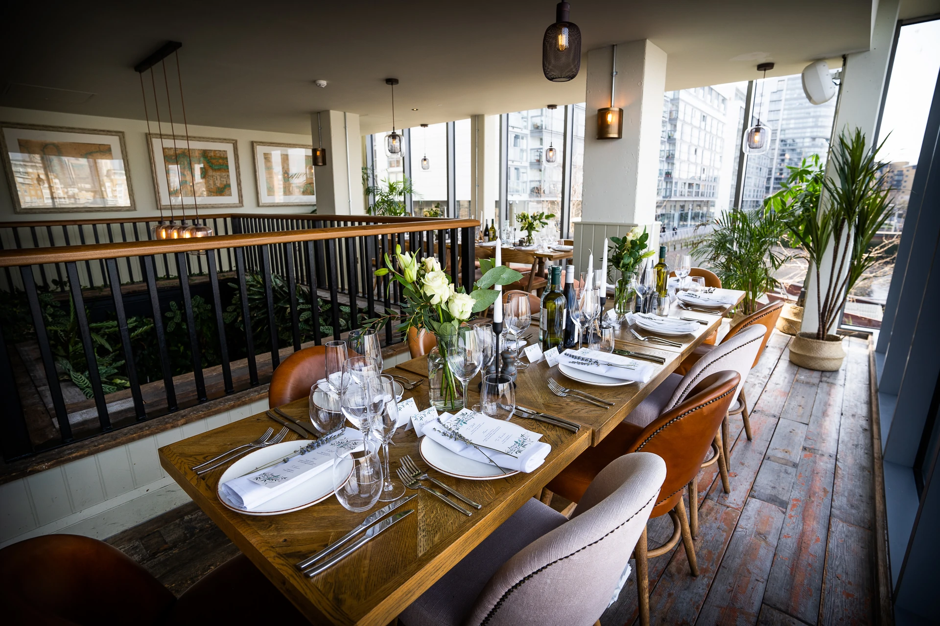 The image depicts a sophisticated restaurant with a large wooden dining table set for six, surrounded by chairs and adorned with white plates, silverware, and flowers. The table is set on a wooden floor, with a black railing and a black railing in the background. The room features framed pictures on the wall, hanging lights, and a large window offering a view of the city. The restaurant is well-lit and offers a cozy atmosphere.