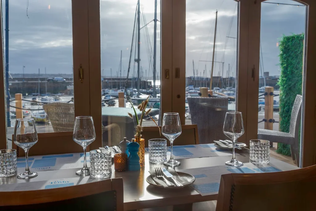 The image features a serene dining area with a wooden table set for two, surrounded by chairs. The table is adorned with a blue tablecloth, silverware, and a vase with a green plant. The background shows a marina with boats, a blue sky, and a window revealing the outside view. The scene is set against a backdrop of a blue sky and white clouds, creating a tranquil atmosphere.