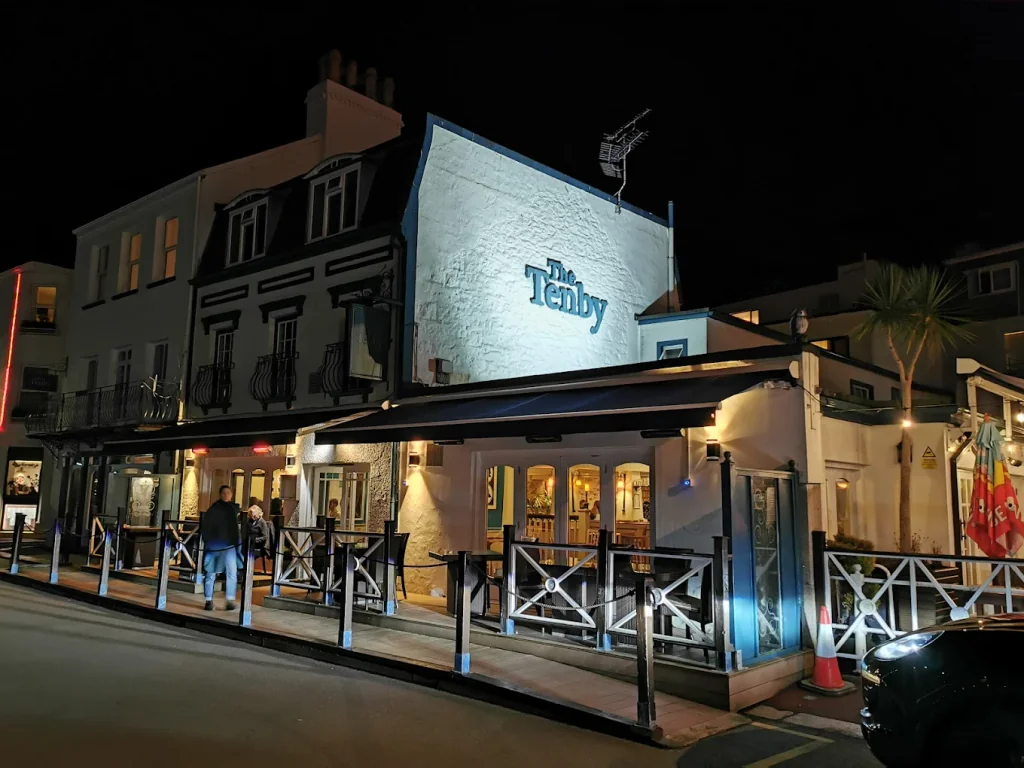 The image features the "The Bluebird" pub in Plymouth, UK, with a white facade, black roof, and blue sign. The photo is taken from a street corner, with a sidewalk in front of the pub and a car parked nearby. The scene is illuminated by warm lights, creating a cozy atmosphere.