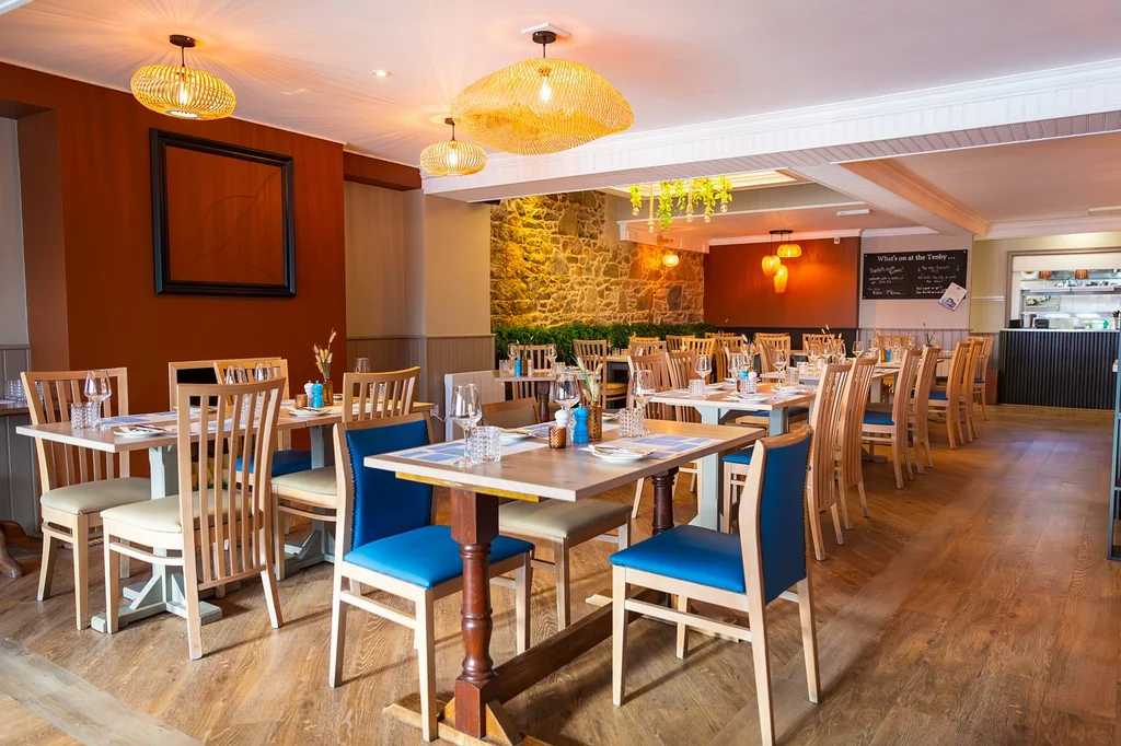 The image shows a modern, well-lit restaurant with wooden floors and walls, featuring wooden chairs and tables with blue and white tablecloths. The tables are set with white plates, silverware, and condiments. Yellow chandeliers hang from the ceiling, casting a warm glow. A stone wall with a blackboard menu is in the background, and a wooden counter is in the foreground. The arrangement of objects creates a harmonious and inviting atmosphere.