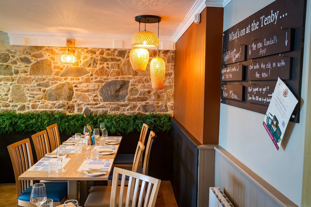The image shows a cozy restaurant with a rustic stone wall, a stone wall with a green hedge, and a wooden table with white plates, silverware, and napkins. Two hanging light fixtures with yellow shades illuminate the space, and a menu board with English and French text is on the wall. The wooden floor and chairs add warmth, and the overall atmosphere is inviting and welcoming.