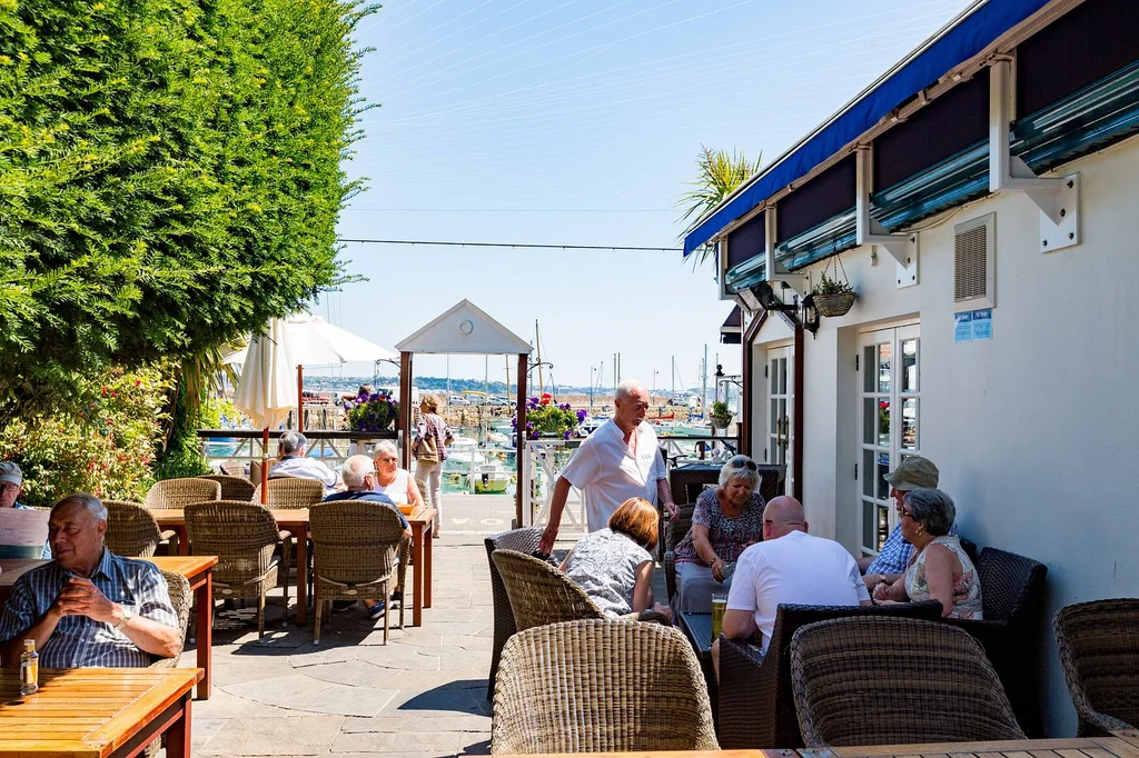 A lively outdoor café on a beachside terrace overlooks the ocean, with people enjoying the view and a man in a white shirt conversing with a woman. The café is surrounded by lush greenery and a white building with a blue awning. The clear blue sky and ocean create a serene atmosphere, making it a perfect spot for relaxation and socializing.