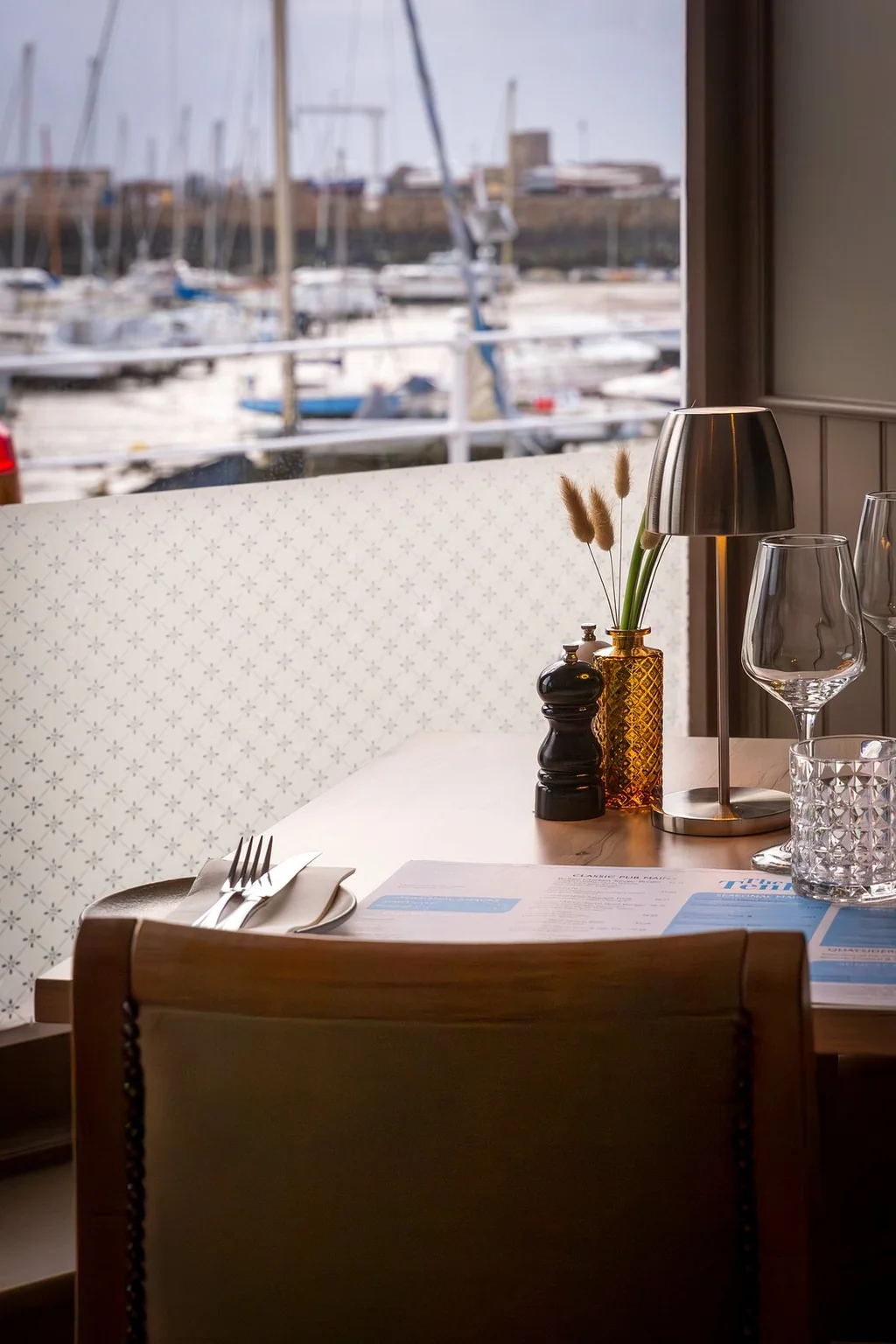 A serene dining scene is captured in a marina view, with a wooden table set for two with a silver fork, knife, and napkin, a vase of flowers, and a lamp. The table is near a window, offering a view of the harbor with boats and a building. The objects are arranged harmoniously, creating a tranquil atmosphere.
