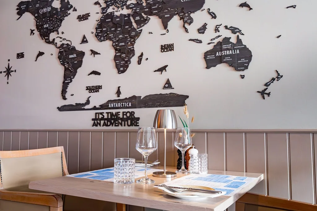A cozy dining room features a white tablecloth with a blue and white checkered napkin, silverware, and wine glasses. A black world map with "African" text is on the wall behind the table. The room is illuminated by soft light, creating a warm and inviting atmosphere.