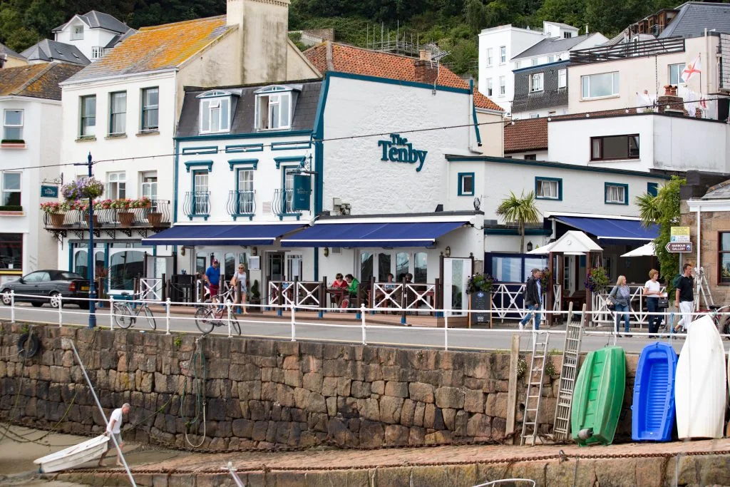 The image depicts a bustling harbor scene with a variety of buildings, including a white one with a blue awning and a blue and white building. The harbor is filled with boats, including a blue one, and people are enjoying the atmosphere. A stone wall separates the harbor from the buildings, and a white fence runs along the harbor. The scene is filled with vibrant colors and details, creating a lively and inviting atmosphere.