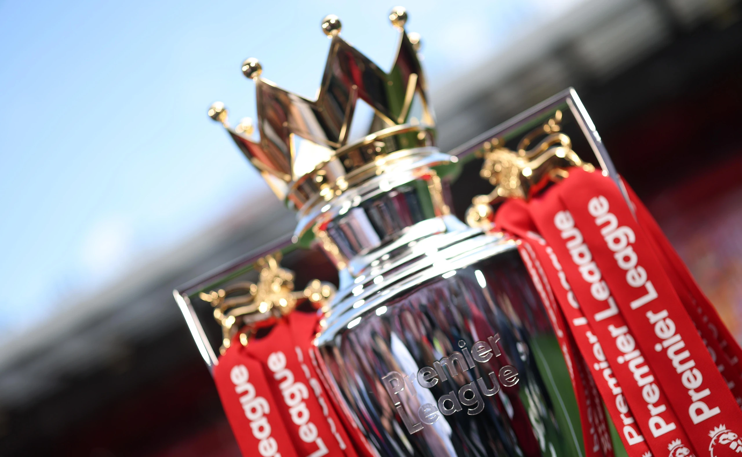 A silver trophy with a gold crown and red banner with "Liverpool" text is the central focus of the image, set against a blurred stadium background. The trophy is the main object, with the banner as a secondary element. The image captures a moment of celebration and achievement, symbolizing the power of the trophy and the significance of the banner.
