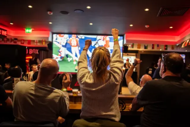 In a lively bar, a group of people gather around a television displaying a football game, cheering and raising their hands. The bar is filled with beer bottles and glasses, and the atmosphere is electric. The red ceiling contrasts with the white walls, and the scene captures a moment of joy and camaraderie.