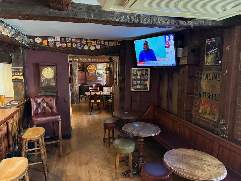 The image shows a cozy pub with rustic charm, featuring wooden floors, a brick wall, and a wooden bar. The bar has stools and a wooden bench, and a television is mounted above. The walls display posters and a clock, and the pub has a warm, inviting atmosphere.