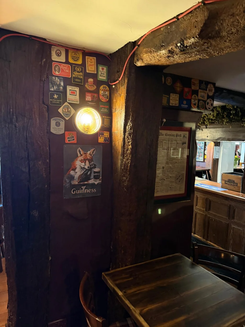 In a dimly lit, rustic pub, a wooden wall adorned with a cat poster and a red rope hangs above a wooden table. A wooden bench is positioned in front of the table, and a menu hangs on the wall. The scene is bathed in warm, inviting light, creating a cozy atmosphere.